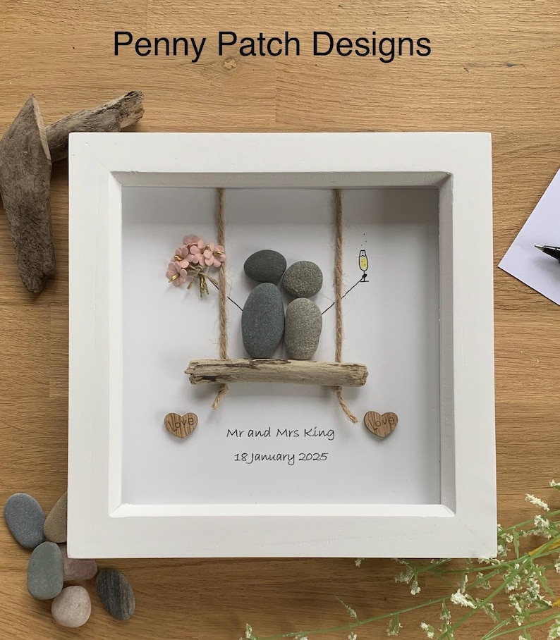 May include: White framed artwork featuring a pebble art scene of a couple on a swing. The swing is made of driftwood and rope, with a small bouquet of pink flowers. The artwork includes the text "Mr and Mrs King 18 January 2025".