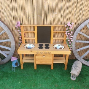 Può includere: Cucina giocattolo in legno con due lavelli, un piano cottura e una lavagna. La cucina è dipinta in un colore legno naturale e ha un aspetto rustico.