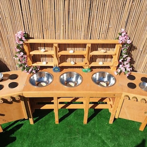 Op de afbeelding: Een houten speelkeuken met een gootsteen, fornuis en planken. De keuken is geschilderd in een natuurlijke houtkleur en heeft een groene gras achtergrond.