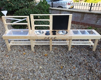 May include: A wooden outdoor play kitchen with two sinks, a stovetop, and a water table. The kitchen has a black chalkboard and shelves for storage.