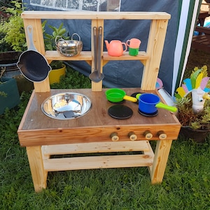 May include: Wooden play kitchen with a stainless steel sink, a stovetop with four knobs, and a shelf for play food and utensils. The kitchen is painted in a natural wood colour and has a small shelf above the stovetop. There are several plastic play kitchen utensils and a unicorn figurine on the counter.