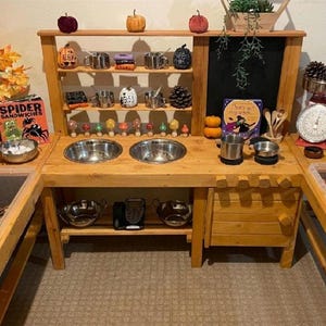 May include: A wooden play kitchen with a black chalkboard, two stainless steel sinks, and a shelf with various Halloween-themed decorations. The kitchen has a clear plastic bin with a spider and pumpkins, a book titled "Spider Sandwiches", and a wooden drawer.