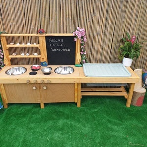 May include: Wooden play kitchen with a chalkboard sign that says "Bella's Little Treasures". The kitchen has two sinks, a shelf, and a storage area. There are also some decorative items, such as a ladybug and flowers.