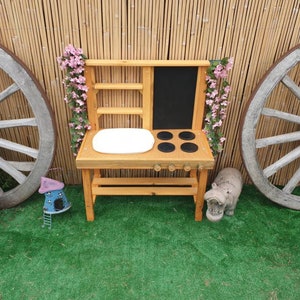 Può includere: Cucina giocattolo in legno con un lavandino bianco, una lavagna nera e un piano cottura a quattro fuochi. La cucina è decorata con ghirlande di fiori rosa. La cucina è installata in un giardino con un prato verde e due ruote di carro di legno.
