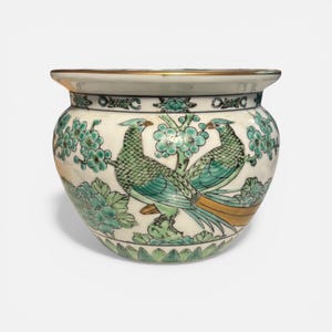 May include: A decorative ceramic planter with a gold rim. The planter features a detailed design of two birds with teal and gold feathers, surrounded by teal floral patterns on a white background.