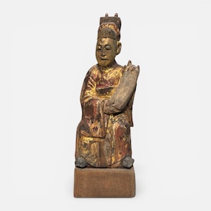 May include: A carved wooden statue of a seated figure with a tall hat, holding a scroll. The statue is painted with gold, red, and brown hues, and is mounted on a rectangular wooden base. The figure's clothing and hat suggest a historical or cultural theme.