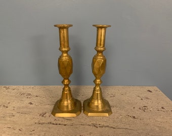 May include: A pair of gold metal candle holders with a geometric design. The candle holders are on a light brown surface.