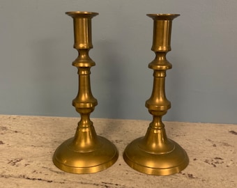 May include: A pair of gold metal candle holders with a classic design. The candle holders have a round base and a tall, slender stem with a decorative knob at the top.