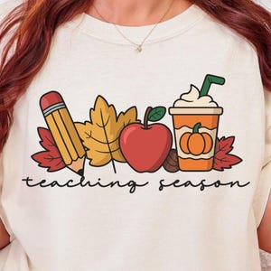 Puede incluir: Camiseta color crema con un diseño temático de otoño. El diseño incluye un lápiz, hojas de otoño, una manzana roja y un café con leche de calabaza. Las palabras "teaching season" están escritas en cursiva.