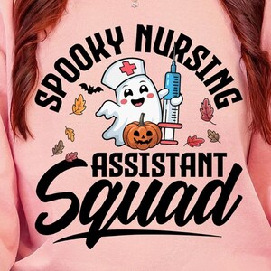 Puede incluir: Una sudadera rosa claro con la frase "Spooky Nursing Assistant Squad" en negro. El diseño incluye un fantasma de dibujos animados con gorro de enfermera, una jeringa y una calabaza. Una taza negra con fantasmas blancos se sostiene en la mano.