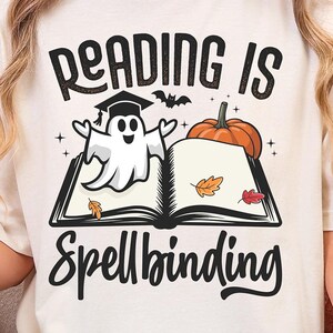 May include: Cream-colored t-shirt with the text "Reading is Spellbinding" in black script. The graphic features a cartoon ghost wearing a graduation cap, a pumpkin, and an open book with autumn leaves. The shirt is a casual, everyday wear item.