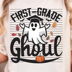 Pode incluir: Camiseta creme com o texto preto "FIRST-GRADE Ghoul", apresentando um fantasma de desenho animado usando um chapéu de formatura, cercado por folhas de outono e uma abóbora. A camiseta é um design temático de Halloween.