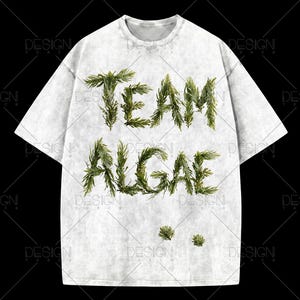 Könnte beinhalten: Ein kurzärmeliges T-Shirt aus hellgrauem, meliertem Stoff mit Acid-Wash-Effekt. Auf der Vorderseite befindet sich der Schriftzug "TEAM ALGAE" in einer dunkelgrünen, strukturierten Schrift, die an Tannennadeln erinnert, mit zwei kleinen grünen Zweigen.