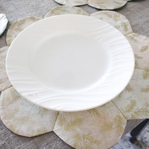 May include: A white ceramic dinner plate with a wavy rim sits on a beige and gold floral placemat. The placemat has a scalloped edge and a pattern of stylized leaves.