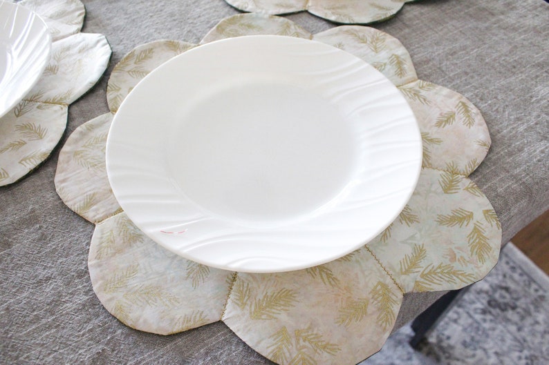 May include: A white ceramic dinner plate with a wavy rim sits on a beige and gold floral placemat. The placemat has a scalloped edge and a pattern of stylized leaves.