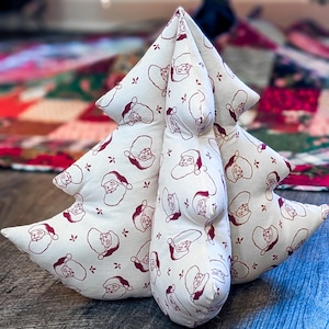 May include: Three Christmas tree-shaped pillows. The pillows are made of off-white fabric with a repeating pattern of Santa Claus faces and small red floral accents. The pillows are arranged to resemble a Christmas tree.