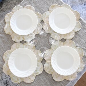 May include: Four white ceramic dinner plates with a raised rim are placed on four beige and gold floral patterned placemats. The placemats have a scalloped edge.