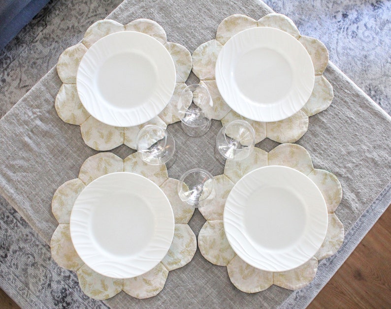 May include: Four white ceramic dinner plates with a raised rim are placed on four beige and gold floral patterned placemats. The placemats have a scalloped edge.