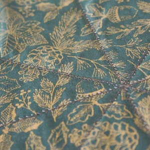 May include: Close-up of a teal fabric with a gold leaf pattern. The fabric is quilted with a brown thread.