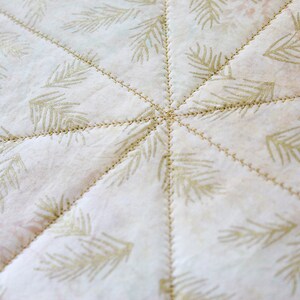 May include: A white fabric with a repeating pattern of gold pine branches. The fabric is quilted with a starburst pattern.