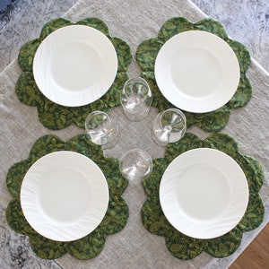 May include: Four white dinner plates are set on green floral placemats. The placemats have a scalloped edge and are arranged on a light gray tablecloth.