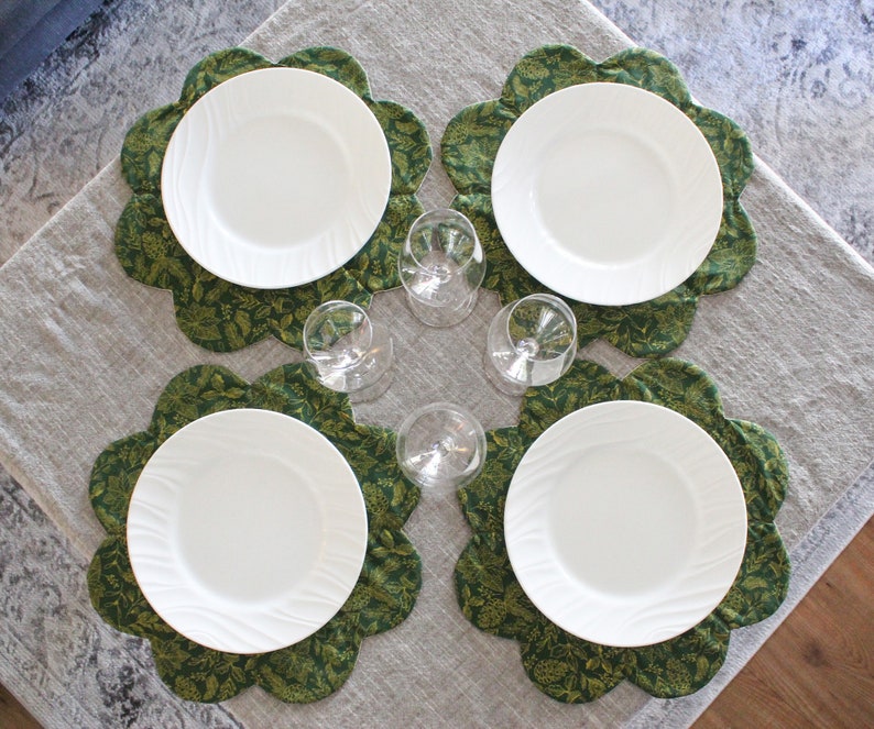 May include: Four white dinner plates are set on green floral placemats. The placemats have a scalloped edge and are arranged on a light gray tablecloth.