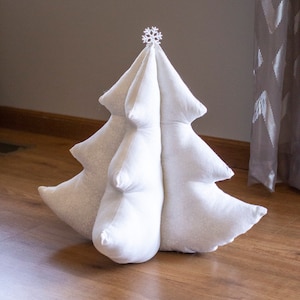 May include: A white, fabric Christmas tree-shaped pillow with a snowflake topper. The tree has a soft, quilted appearance with a subtle, repeating pattern. The pillow is standing on a wooden floor.