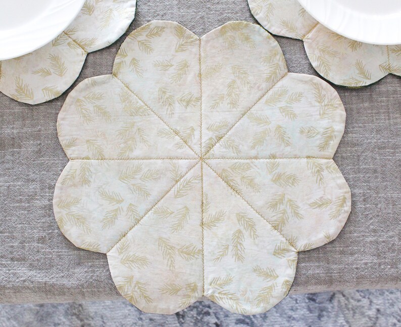May include: A white fabric placemat with a gold leaf pattern. The placemat is shaped like a flower with six points.
