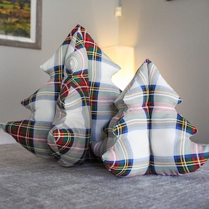 May include: Two plaid Christmas tree pillows, one larger than the other, are sitting on a gray linen surface. The pillows are made of a white fabric with a red, green, blue, and yellow plaid pattern.
