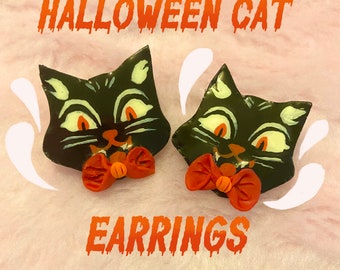 May include: A pair of Halloween cat earrings. Each earring features a black cat with a red bow tie and white eyes. The text "HALLOWEEN CAT EARRINGS" is printed in dripping red letters.