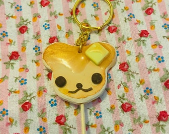 May include: A gold and brown bear-shaped pancake on a stick with a yellow square of butter on top. The pancake is attached to a gold key chain.