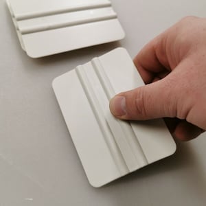 May include: Two off-white plastic squeegees, one held by a hand. Each squeegee has a rectangular shape with rounded corners and three parallel grooves. The squeegees are designed for smoothing surfaces.