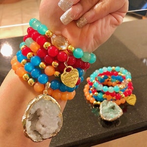 May include: A stack of colorful beaded bracelets with gold accents and a large, white, crystal-shaped pendant. The bracelets feature turquoise, red, orange, and gold beads. The pendant is attached to a gold chain.