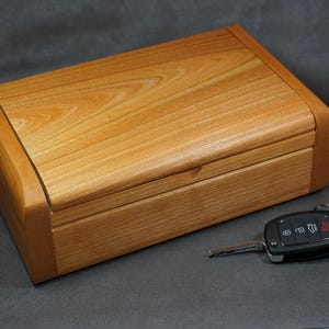 May include: A wooden box with a hinged lid, made of light brown wood with a natural wood grain. The box is closed and a car key is lying next to it.