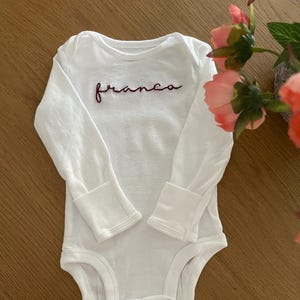 Op de afbeelding: Witte babybody met lange mouwen en een ronde halslijn. Het woord "franco" is in sierlijke letters in donkerrood op de borst geborduurd. De body heeft drukknoopsluitingen aan de onderkant.