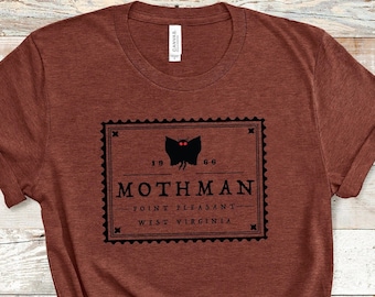 May include: A brown t-shirt with a black and white graphic of a mothman with red eyes. The graphic is in the shape of a postage stamp with the text "Mothman" and "Point Pleasant, West Virginia".