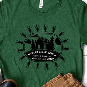 May include: A green t-shirt with a black graphic of a cabin and the text "Winter River Bridge, Winter River, CT, Come back again soon!" surrounded by a circle of black beetle silhouettes.