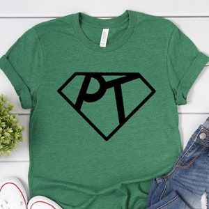 May include: A green t-shirt with a black diamond-shaped graphic that says "PT" inside. The shirt is folded and displayed on a white surface with a pair of white sneakers and blue jeans.