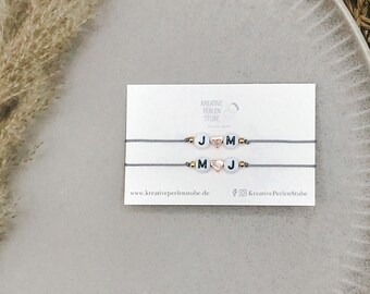 May include: Two grey string bracelets on a white display card labelled Kreative Perlen Stube. Each bracelet features white round beads with black letters J and M, separated by a small rose gold heart bead and accented with tiny metallic gold beads.