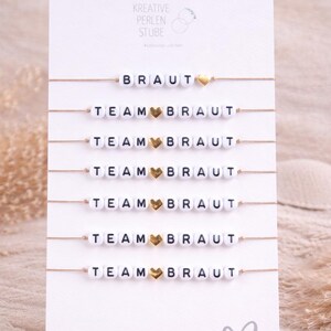 Op de afbeelding: Een set van zeven dunne armbanden op een witte displaykaart. Eén armband bevat de tekst BRAUT met een gouden hartje, en zes armbanden bevatten de tekst TEAM BRAUT met een gouden hartje. De kralen zijn wit met zwarte letters en gouden accenten.