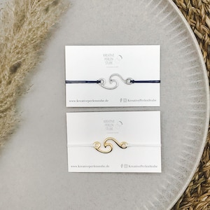 Può includere: Due braccialetti su cartoncini bianchi. Il braccialetto superiore ha un ciondolo a forma di onda color argento su un cordino blu scuro. Quello inferiore ha un ciondolo a forma di onda color oro su un cordino bianco. Ogni cartoncino riporta il testo KREATIVE PERLEN STUBE.