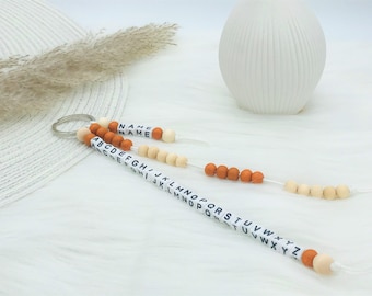 以下が含まれることがあります： 金属製のリングに3本のビーズの紐が付いたキーホルダー。1本の紐には白いキューブに黒い文字でNAMEと書かれています。他の2本の紐には白いキューブにAからZまでのアルファベットが記されており、オレンジとベージュの木製ビーズが交互に配置されています。