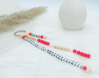 以下が含まれることがあります： 金属製のリングに2本の白いコードが付いたキーホルダー。一方のコードには白いビーズに黒い文字でNAMEと書かれており、もう一方のコードには白いビーズに黒い文字でAからZまでのアルファベットがすべて記されています。赤とベージュのビーズがアクセントになっています。