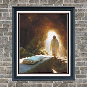 Puede incluir: Una impresión enmarcada representa una figura con una túnica blanca saliendo de una cueva iluminada. Una losa de piedra con una tela blanca está en primer plano. La obra de arte está enmarcada en un borde negro.