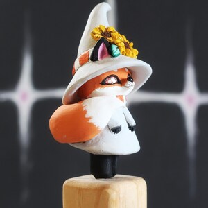 May include: A charming fox figurine, wearing a white witch's hat decorated with yellow sunflowers and a scarf. The fox has orange fur and a white face, mounted on a wooden base. The figurine is approximately 10 cm tall.