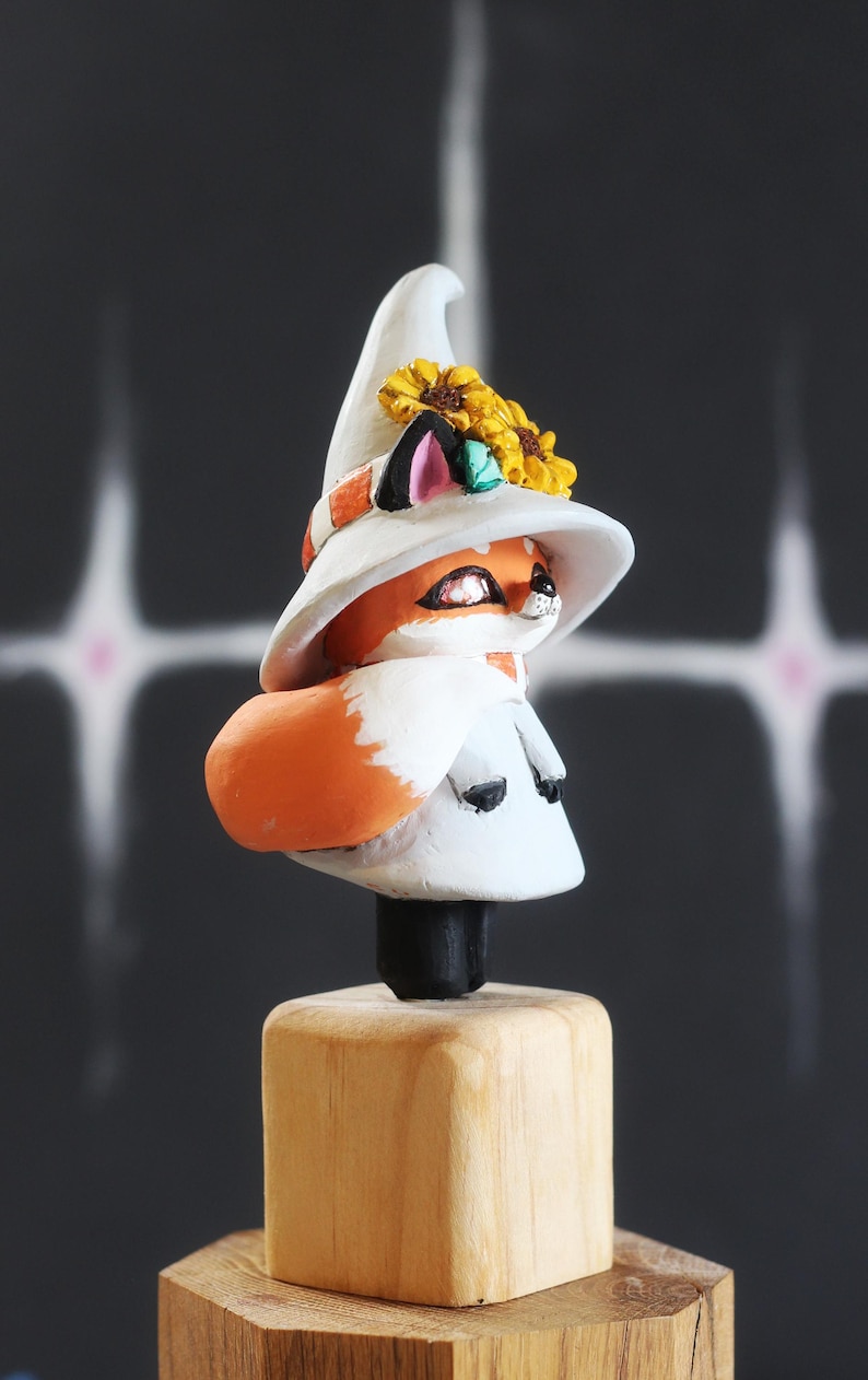 May include: A charming fox figurine, wearing a white witch's hat decorated with yellow sunflowers and a scarf. The fox has orange fur and a white face, mounted on a wooden base. The figurine is approximately 10 cm tall.