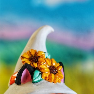 May include: A decorative fox figurine wearing a white witch's hat decorated with two yellow sunflowers. The fox has orange fur, pink ears, and a striped scarf. The background is a blurred mix of blue, green, and pink.