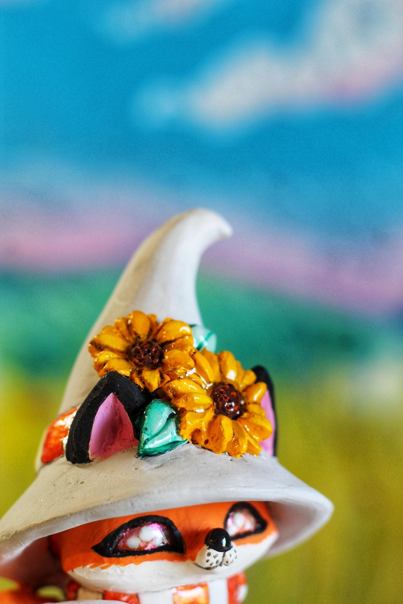 May include: A decorative fox figurine wearing a white witch's hat decorated with two yellow sunflowers. The fox has orange fur, pink ears, and a striped scarf. The background is a blurred mix of blue, green, and pink.