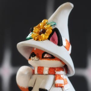 May include: A decorative fox figurine, featuring a white witch's hat with a sunflower, and an orange and white scarf. The fox has orange fur, black ears, and a white coat. A charming, whimsical design.