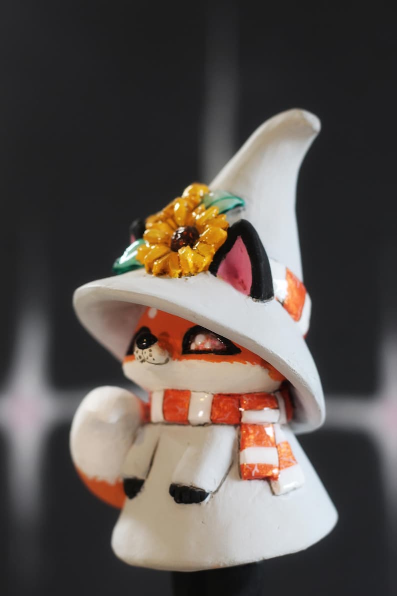 May include: A decorative fox figurine, featuring a white witch's hat with a sunflower, and an orange and white scarf. The fox has orange fur, black ears, and a white coat. A charming, whimsical design.